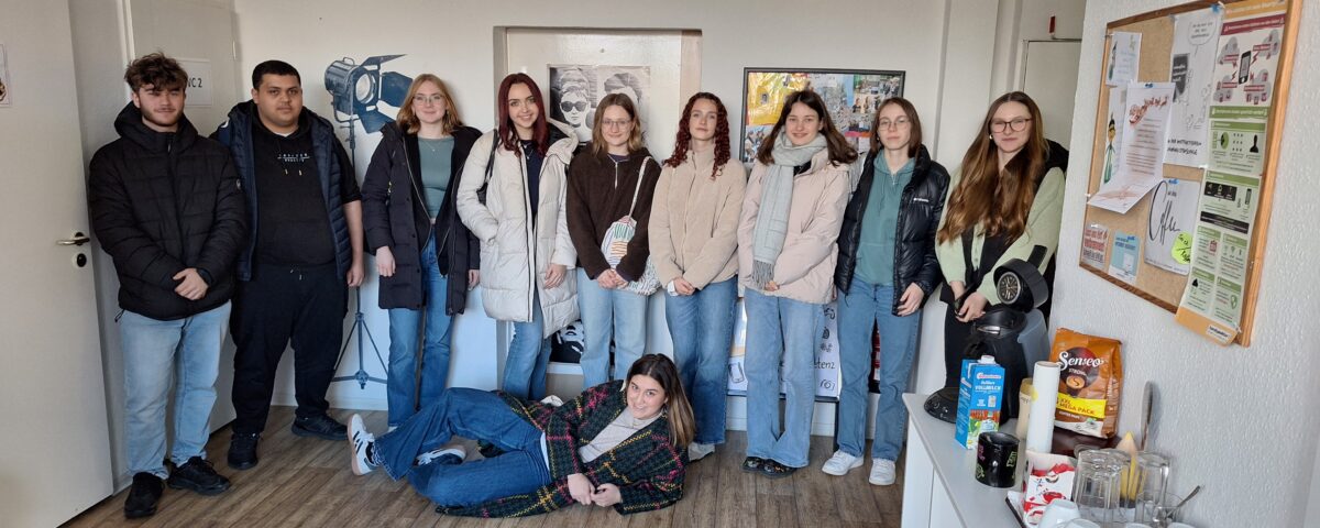 Das Bild zeigt eine Gruppe von elf Schülern in einem Raum mit Holzboden und wei-ßen Wänden. Zehn Personen stehen und eine liegt. Die meisten Personen tragen winterliche Kleidung. Die Gruppe ist gemischt, mit mehreren Frauen und zwei Män-nern auf der linken Seite. Die Gruppe steht entspannt zusammen, einige lächeln leicht. Die Person in der Mitte des Vordergrunds liegt auf dem Boden und stützt sich auf einen Ellenbogen. Die Stimmung wirkt locker und freundlich.